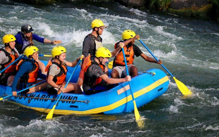 A group of students wearing safety gear paddle a raft through whitewater. 